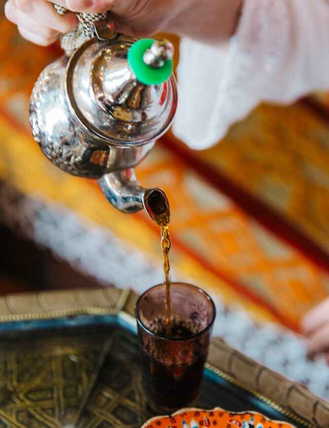 serving moroccan mint tea Moroccan mint tea teapot pouring mint tea from a height into a small glass on a wooden table, with foam forming at the top